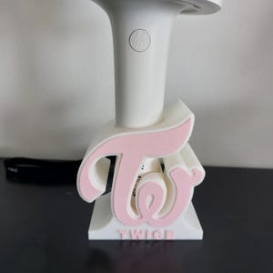 Könnte beinhalten: Ein weißer Leuchtstab mit einem rosa-weißen Ständer. Der Ständer zeigt das Wort "TWICE" in Rosa, wobei das "T" und das "W" ein stilisiertes Design bilden. Der Leuchtstab wird in den Ständer eingesetzt.