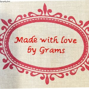 May include: A red oval frame with a decorative border on a beige fabric background. The text inside the frame reads "Made with love by Grams".