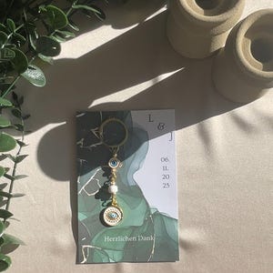 May include: A gold-colored keychain with an evil eye charm and white beads, displayed on a card with the text "Herzlichen Dank". The card has a green and white marble design. The keychain is a decorative accessory.