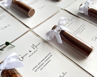 May include: Arrangement of wedding favors featuring small glass vials filled with a brown substance, tied with white ribbons. The vials rest on white cards with printed text and names.