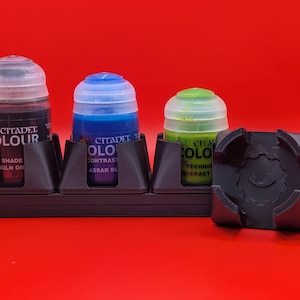 Op de afbeelding: Een donkerbruine 3D-geprinte verfpotjeshouder met drie Citadel Colour verfpotjes en een aparte houder. De potjes bevatten rode, blauwe en groene verf. De tekst "Citadel Colour" is zichtbaar op de potjes.
