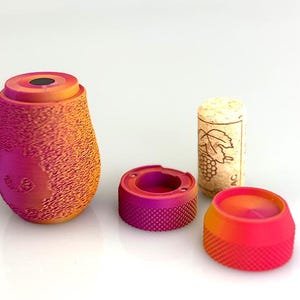 May include: A collection of wine-related items, including a textured, gradient-colored bottle stopper, two matching rings, and a cork. The stopper and rings feature a textured surface and a color gradient from orange to pink. The cork has a grape design.