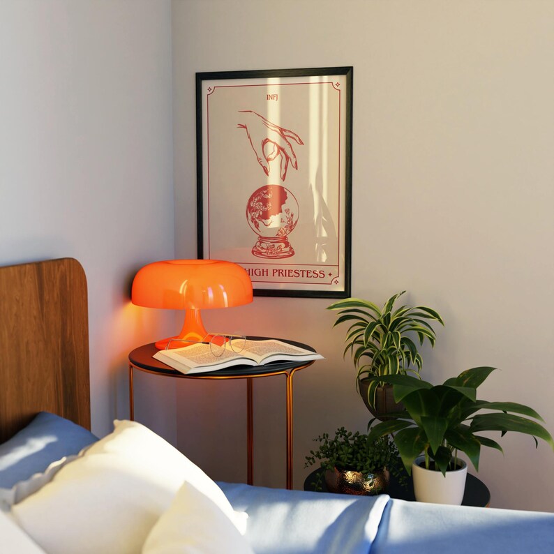 May include: An orange mushroom-shaped lamp sits on a black side table next to a bed. A framed print with a hand over a crystal ball is on the wall. A book and glasses are on the table. Plants in pots are on the table.