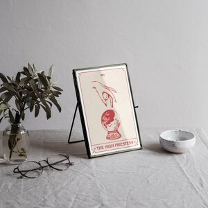 May include: A framed print of "The High Priestess" tarot card. The artwork features a hand reaching towards a crystal ball, all in red on a cream background. The frame is green with a black stand. A small white bowl and a vase with greenery are also present.