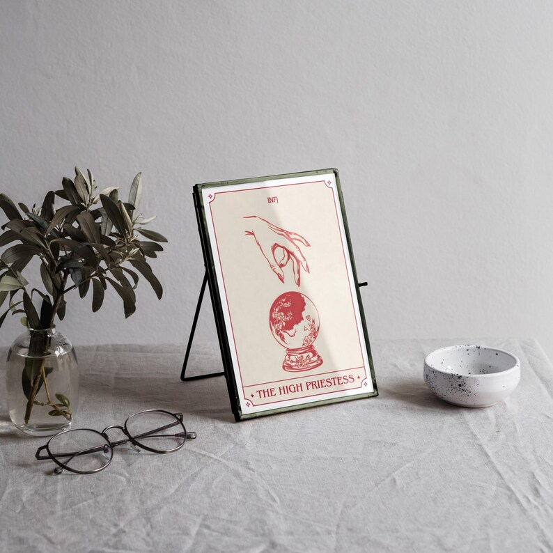May include: A framed print of "The High Priestess" tarot card. The artwork features a hand reaching towards a crystal ball, all in red on a cream background. The frame is green with a black stand. A small white bowl and a vase with greenery are also present.
