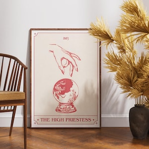 May include: A framed print featuring a red and beige design. The artwork depicts a hand reaching towards a crystal ball, with the text "INFJ" at the top and "THE HIGH PRIESTESS" at the bottom. The print is displayed on a wooden floor.
