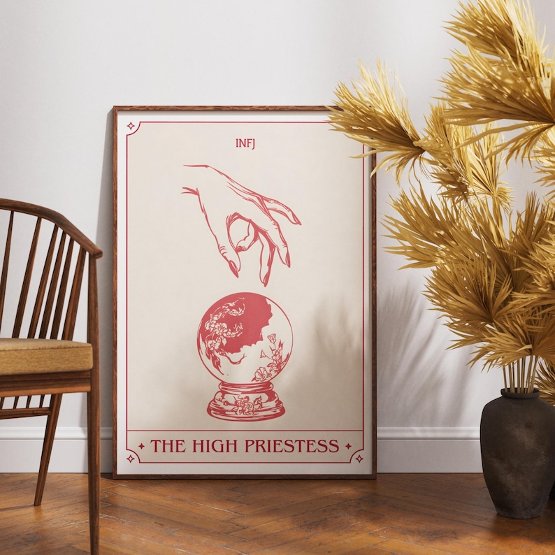 May include: A framed print featuring a red and beige design. The artwork depicts a hand reaching towards a crystal ball, with the text "INFJ" at the top and "THE HIGH PRIESTESS" at the bottom. The print is displayed on a wooden floor.