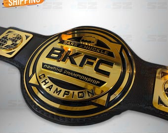Puede incluir: Un cinturón de campeonato con una gran placa metálica circular de color dorado con detalles en negro. La placa muestra el texto BARE KNUCKLE BKFC FIGHTING CHAMPIONSHIP CHAMPION en relieve. Una correa de cuero negro se extiende desde la placa central.