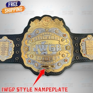 Puede incluir: Un cinturón de campeonato dorado y plateado con el texto "HEAVYWEIGHT" y "IWGP CHAMPION". El cinturón tiene una correa de cuero negro y placas laterales decorativas. La imagen también incluye el texto "FREE SHIPPING".
