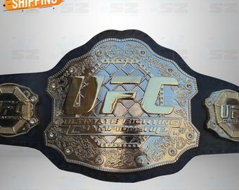May include: A black leather championship belt featuring a large, central gold-toned metal plate with the text UFC ULTIMATE FIGHTING CHAMPIONSHIP embossed in the centre. The plate has a chain-link pattern background and two smaller side plates.