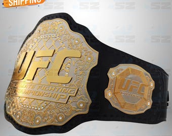 May include: A black championship belt featuring a large, ornate silver and gold central plate with the text UFC ULTIMATE FIGHTING CHAMPIONSHIP and a smaller side plate with the text UFC CHAMPION, both accented with intricate metal detailing.