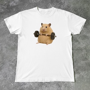 Könnte beinhalten: Ein weißes T-Shirt mit kurzen Ärmeln und einem mittigen Aufdruck eines hellbraunen Hamsters, der eine dunkelgraue Hantel hebt. Der Hamster hat kleine Ohren, schwarze Augen und eine rosa Nase und ist in sitzender Position mit dem Gewicht abgebildet.