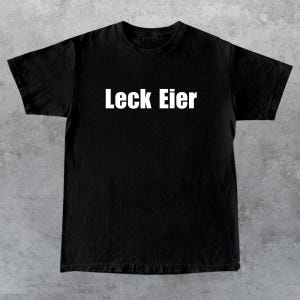 May include: Black t-shirt with the white text "Leck Eier" across the chest. The shirt is made of a soft material and has short sleeves and a crew neck. The background is a textured gray surface.