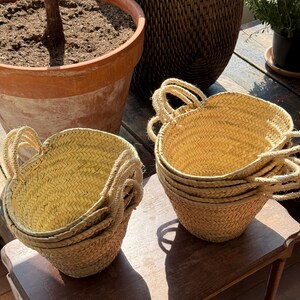 May include: Several small, woven straw baskets with rope handles are stacked on a wooden table. The baskets are a light tan color and have a rounded shape. A large terracotta pot and a woven basket are in the background.