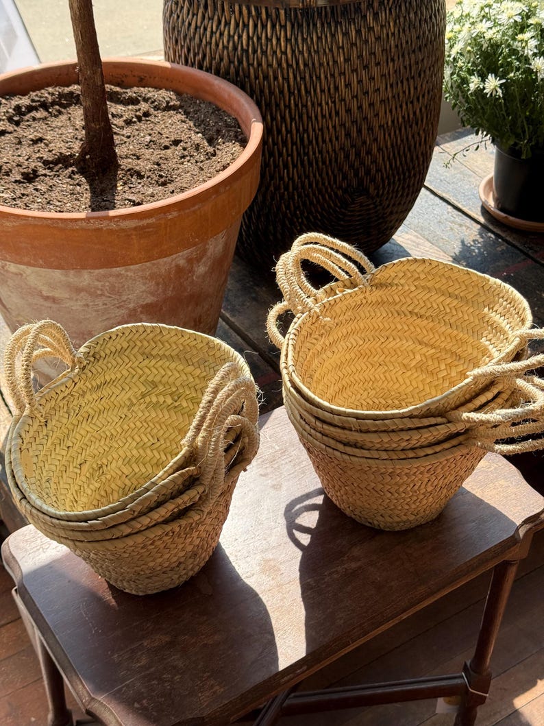 May include: Several small, woven straw baskets with rope handles are stacked on a wooden table. The baskets are a light tan color and have a rounded shape. A large terracotta pot and a woven basket are in the background.