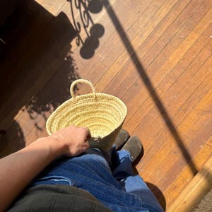 May include: A small, round, woven straw basket with a handle. The basket is held by a person wearing blue jeans and a dark green shirt. The basket is sitting on a wooden floor.