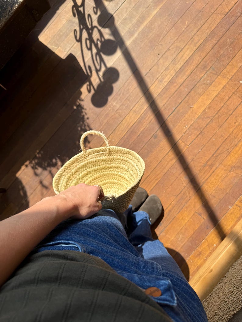 May include: A small, round, woven straw basket with a handle. The basket is held by a person wearing blue jeans and a dark green shirt. The basket is sitting on a wooden floor.