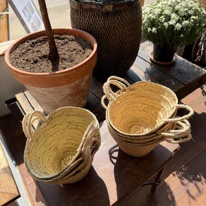 May include: A collection of woven straw baskets with rope handles, stacked on a wooden table. A large terracotta pot with a plant and a dark wicker storage container are in the background. A small potted plant with white flowers is also visible.