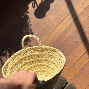 May include: A woven straw basket with a rounded shape and a handle. The basket is held by a person, with the focus on the basket's texture and design. The background shows a wooden floor and shadows.