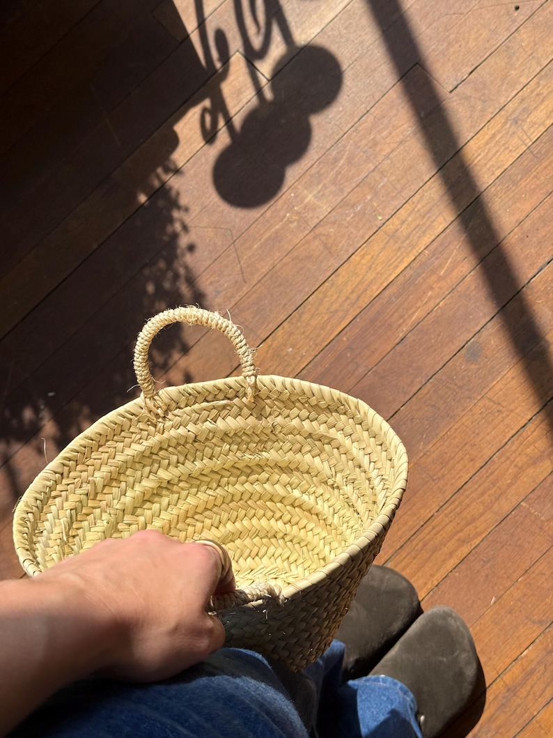 May include: A woven straw basket with a rounded shape and a handle. The basket is held by a person, with the focus on the basket's texture and design. The background shows a wooden floor and shadows.
