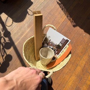 May include: A woven straw basket with a handle, containing a small ceramic mug, a postcard with a building image, a brown box, and an orange leather strap. The basket is held by a person wearing blue jeans.