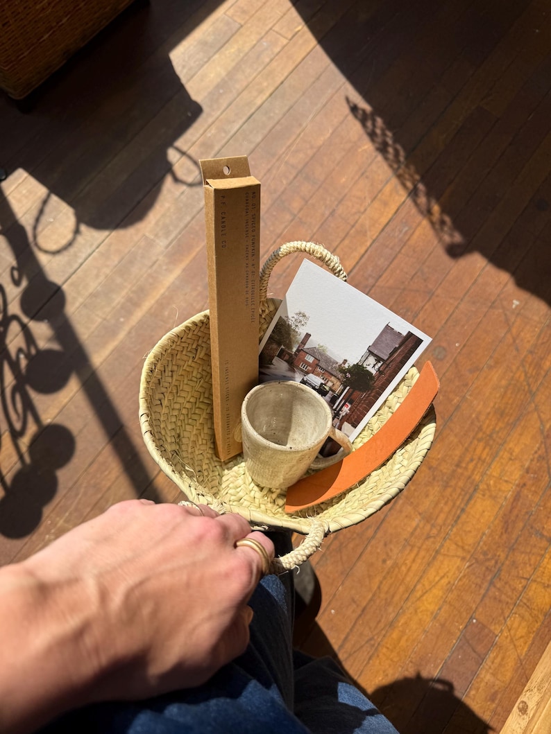 May include: A woven straw basket with a handle, containing a small ceramic mug, a postcard with a building image, a brown box, and an orange leather strap. The basket is held by a person wearing blue jeans.