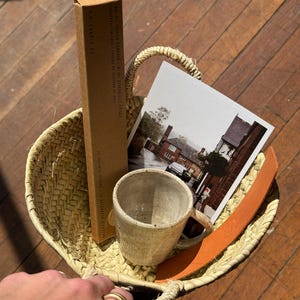 May include: A woven straw basket holds a ceramic mug, a postcard, a brown leather strap, and a box of incense sticks. The box reads "F. CANDLE CO." and "NO. 01 SPICED PUMPKIN". The basket is on a wooden surface.