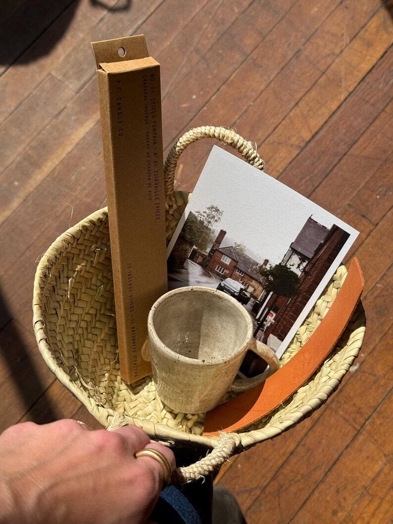 May include: A woven straw basket holds a ceramic mug, a postcard, a brown leather strap, and a box of incense sticks. The box reads "F. CANDLE CO." and "NO. 01 SPICED PUMPKIN". The basket is on a wooden surface.