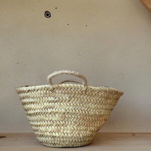 May include: A small, woven basket with two rope handles. The basket is made of light-colored straw and has a flared shape. It sits on a wooden shelf against a neutral-toned wall. The basket's texture and design suggest a rustic, natural aesthetic.