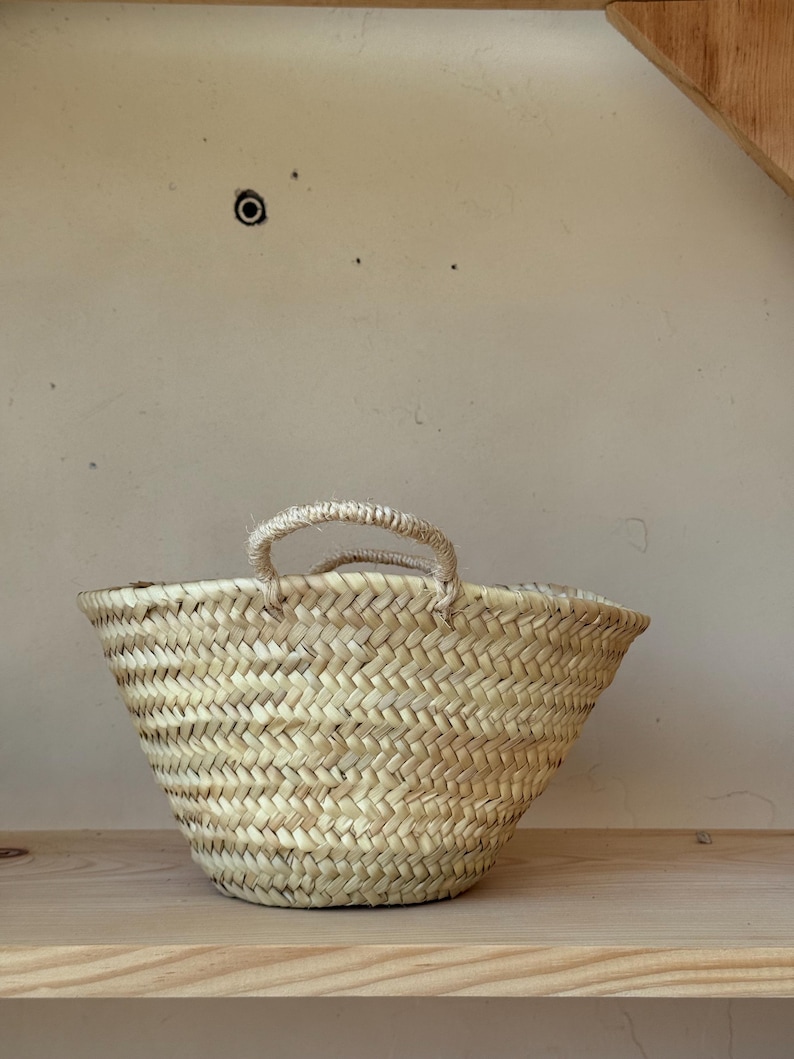 May include: A small, woven basket with two rope handles. The basket is made of light-colored straw and has a flared shape. It sits on a wooden shelf against a neutral-toned wall. The basket's texture and design suggest a rustic, natural aesthetic.
