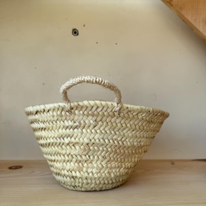 May include: A small, woven basket with a single handle. The basket is made of natural-colored straw and has a rounded shape. It sits on a light-colored wooden shelf, with a neutral background.