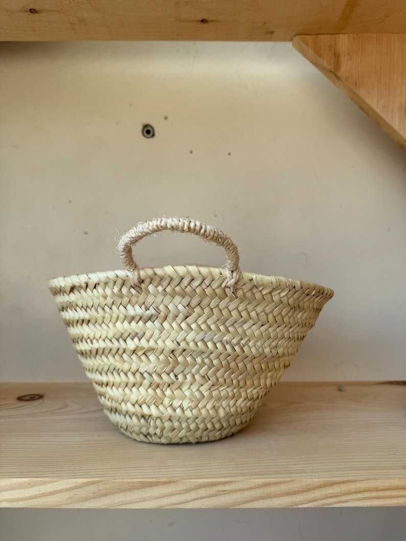 May include: A small, woven basket with a single handle. The basket is made of natural-colored straw and has a rounded shape. It sits on a light-colored wooden shelf, with a neutral background.
