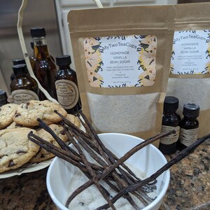 Op de afbeelding: Een assortiment vanilleproducten, waaronder vanillestokjessuiker in kraftpapieren zakken, vanille-extract in kleine glazen flesjes, vanillestokjes en chocolate chip cookies. De zakken zijn voorzien van het label "Homemade Vanilla Bean Sugar".
