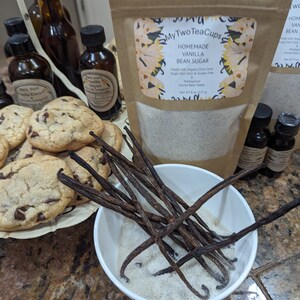 Op de afbeelding: Een presentatie van zelfgemaakte vanilleproducten, waaronder vanillestokjessuiker, extract en koekjes. Een witte kom bevat vanillestokjessuiker en donkerbruine vanillestokjes. Flessen extract en een zak suiker zijn ook zichtbaar.
