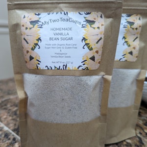Op de afbeelding: Twee bruine papieren zakjes met zelfgemaakte vanillesuiker. Elk zakje heeft een doorzichtig venster en een etiket met de tekst "My Two Tea Cups", "Homemade Vanilla Bean Sugar" en andere details. Het nettogewicht is 227 g.