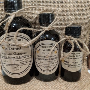 May include: Three glass bottles of vanilla extract, each with a brown paper label and tied with twine. The labels read "100% Pure Vanilla Extract Made with Madagascar Vanilla Beans & Bourbon" and include the maker's name, website, and date. The bottles are arranged from largest to smallest, with the largest bottle being 4 fl. oz., the middle bottle being 2 fl. oz., and the smallest bottle being 1 fl. oz.