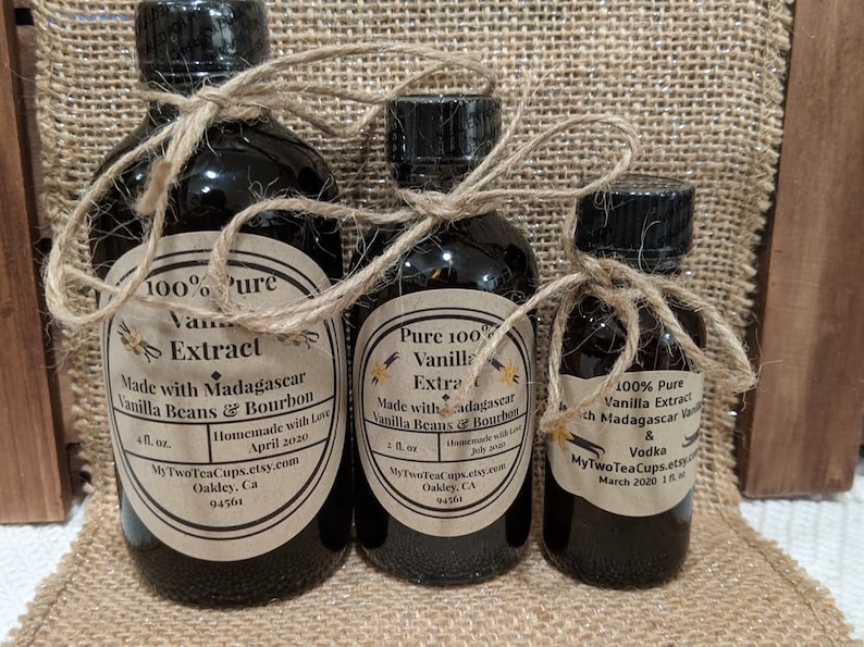 May include: Three glass bottles of vanilla extract, each with a brown paper label and tied with twine. The labels read "100% Pure Vanilla Extract Made with Madagascar Vanilla Beans & Bourbon" and include the maker's name, website, and date. The bottles are arranged from largest to smallest, with the largest bottle being 4 fl. oz., the middle bottle being 2 fl. oz., and the smallest bottle being 1 fl. oz.