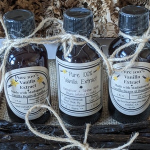 May include: Three glass bottles of pure vanilla extract, each with a brown paper label. The labels feature a yellow flower graphic and the text "Pure 100% Vanilla Extract". The bottles are tied together with twine.