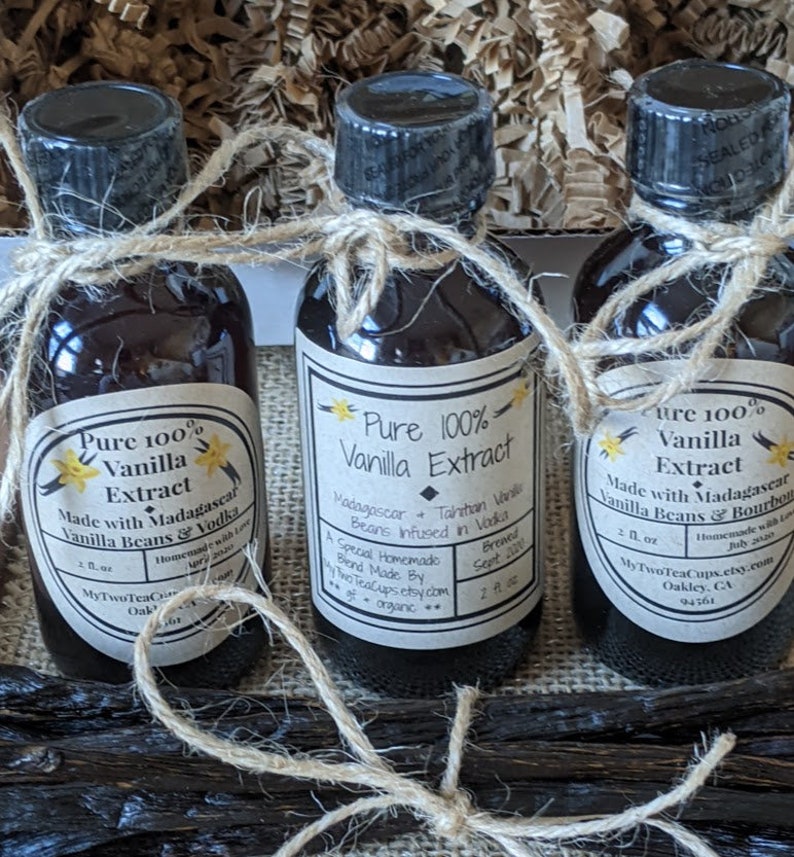May include: Three glass bottles of pure vanilla extract, each with a brown paper label. The labels feature a yellow flower graphic and the text "Pure 100% Vanilla Extract". The bottles are tied together with twine.