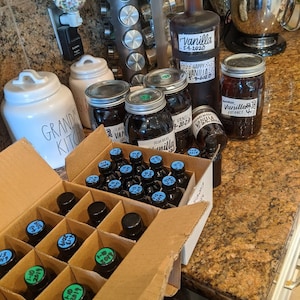 May include: A cardboard box filled with small glass bottles of vanilla extract. The bottles are labeled with dates and the words "Vanilla Extract". There are also several larger glass jars of vanilla extract in the background.