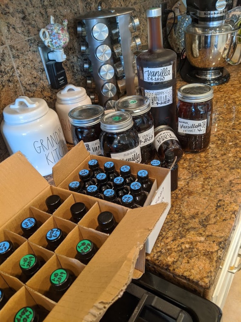 May include: A cardboard box filled with small glass bottles of vanilla extract. The bottles are labeled with dates and the words "Vanilla Extract". There are also several larger glass jars of vanilla extract in the background.