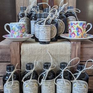 May include: A collection of 10 small glass bottles filled with dark liquid, labeled as "100% Pure Vanilla Extract". The bottles are tied with twine and arranged on a wooden surface with two teacups and saucers.