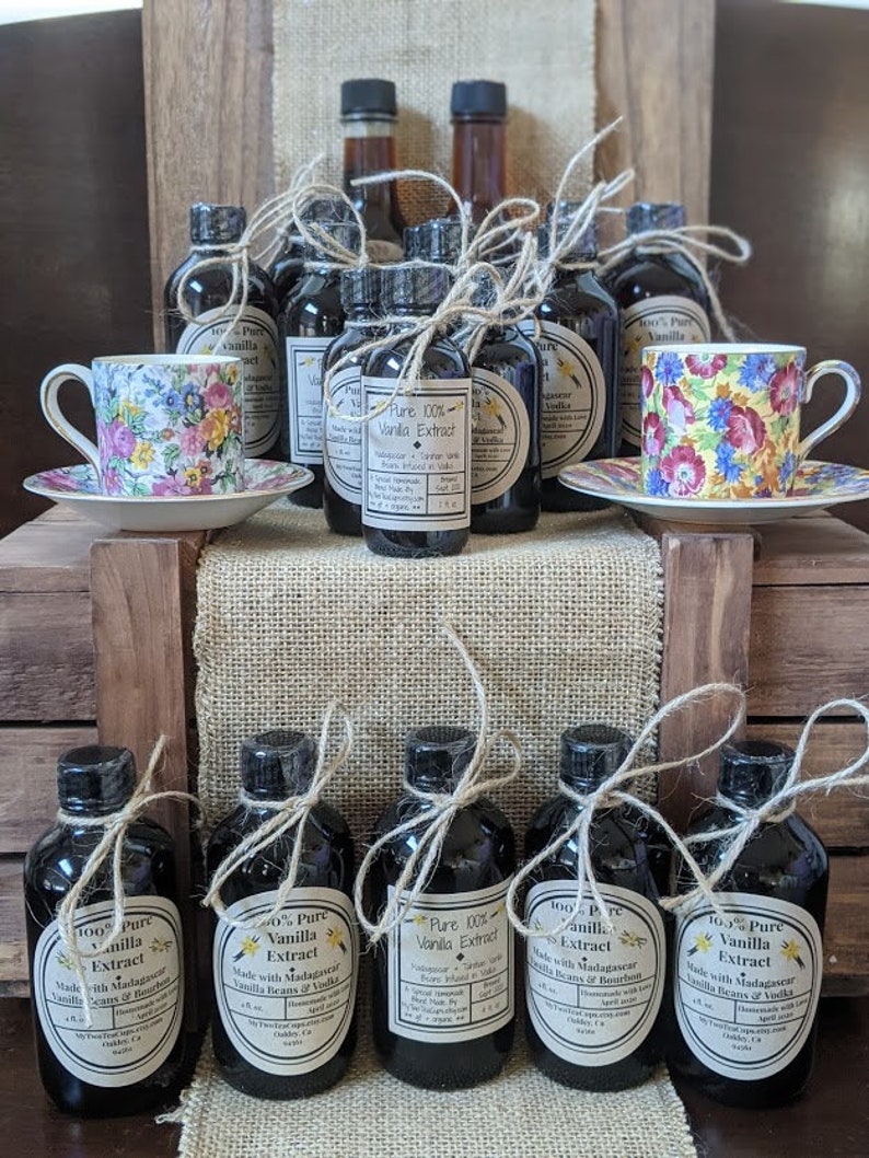 May include: A collection of 10 small glass bottles filled with dark liquid, labeled as "100% Pure Vanilla Extract". The bottles are tied with twine and arranged on a wooden surface with two teacups and saucers.