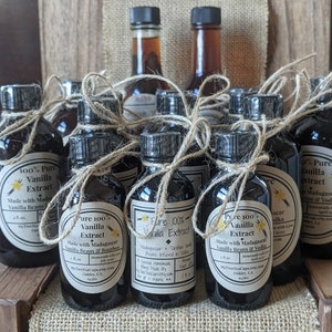 May include: A collection of small glass bottles filled with dark liquid, labeled as "Pure 100% Vanilla Extract". The bottles are tied with twine and arranged on a burlap surface. The labels feature details about the vanilla beans and origin, including "Madagascar" and "Bourbon".