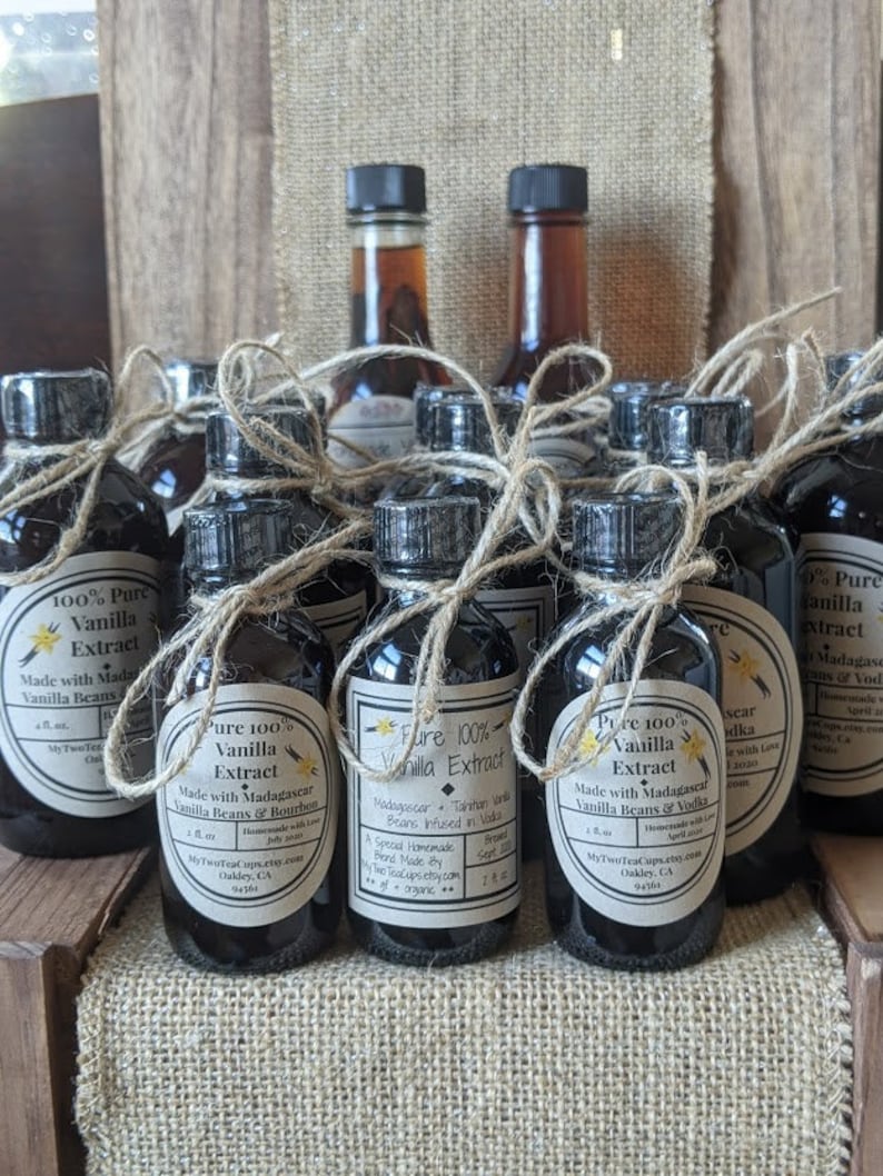 May include: A collection of small glass bottles filled with dark liquid, labeled as "Pure 100% Vanilla Extract". The bottles are tied with twine and arranged on a burlap surface. The labels feature details about the vanilla beans and origin, including "Madagascar" and "Bourbon".