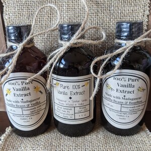 May include: Three glass bottles of pure vanilla extract, each with a different label. The labels are white with black text and feature different types of vanilla beans and alcohol used in the extract. The bottles are tied with twine and are sitting on a burlap surface.