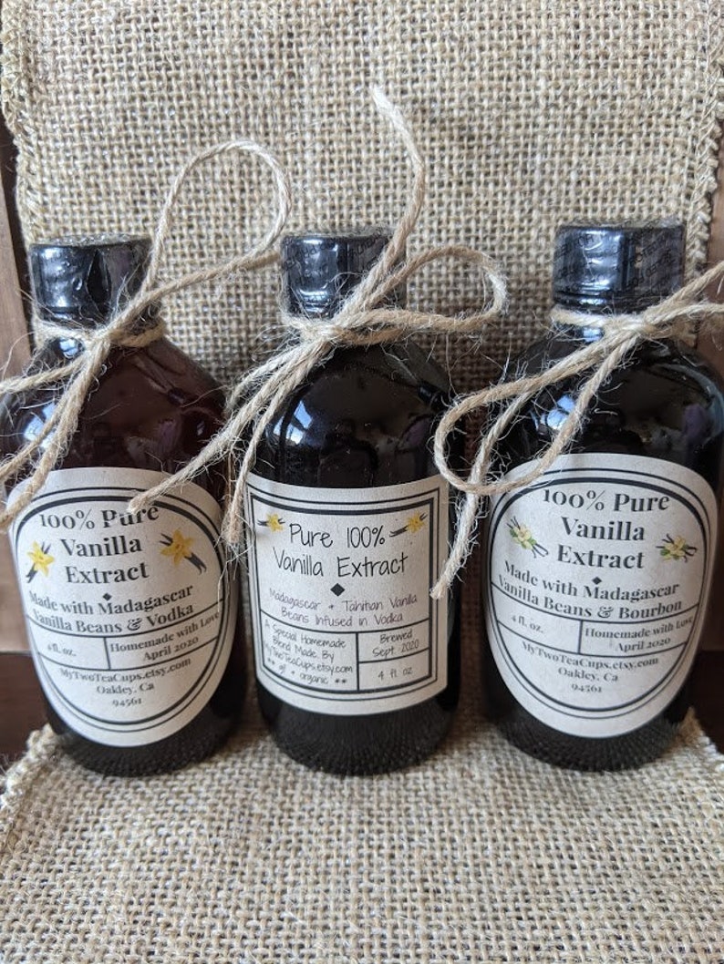 May include: Three glass bottles of pure vanilla extract, each with a different label. The labels are white with black text and feature different types of vanilla beans and alcohol used in the extract. The bottles are tied with twine and are sitting on a burlap surface.