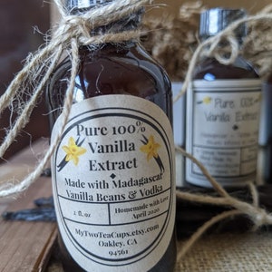 May include: A glass bottle with a brown label that reads "Pure 100% Vanilla Extract Made with Madagascar Vanilla Beans & Vodka Homemade with Love April 2020 MyTwoTeaCups.etsy.com Oakley, CA 94561". The bottle is tied with twine and has a yellow flower graphic on the label.
