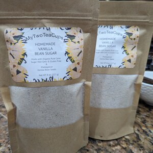 Op de afbeelding: Twee bruine papieren zakken met zelfgemaakte vanille suiker. Elke zak heeft een etiket met de tekst "My Two Tea Cups" en "Homemade Vanilla Bean Sugar". Het etiket vermeldt ook dat de suiker is gemaakt met biologische rietsuiker, niet-GMO, glutenvrij en Madagascar vanillebonen bevat. Elke zak weegt 227 g.