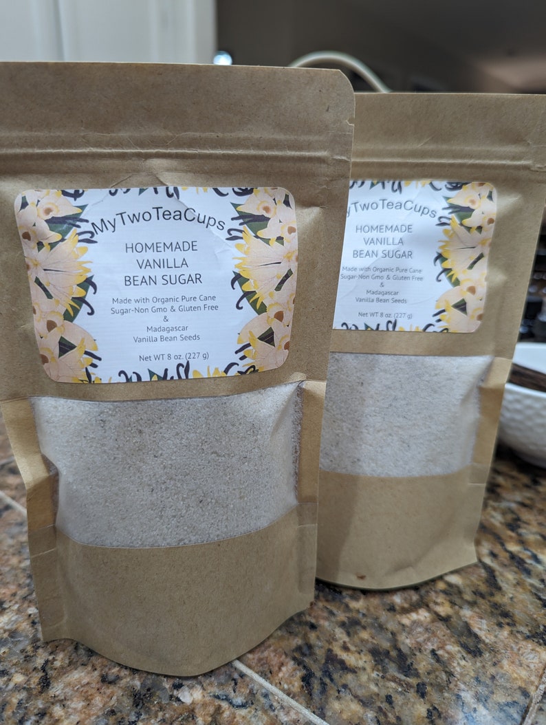 Op de afbeelding: Twee bruine papieren zakken met zelfgemaakte vanille suiker. Elke zak heeft een etiket met de tekst "My Two Tea Cups" en "Homemade Vanilla Bean Sugar". Het etiket vermeldt ook dat de suiker is gemaakt met biologische rietsuiker, niet-GMO, glutenvrij en Madagascar vanillebonen bevat. Elke zak weegt 227 g.